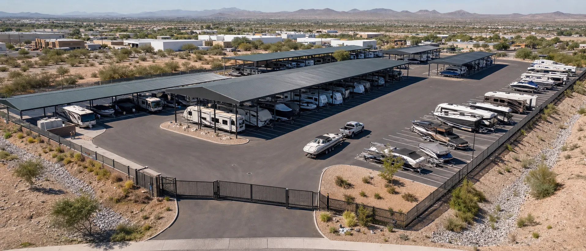 Aerial view of a modern RV and boat storage facility with covered rows, open parking, a gated entrance, broad drive aisles, and desert drainage landscaping.